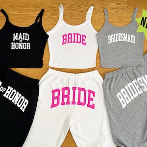 May include: A collection of matching tank tops and sweatpants sets in black, white, and gray. The tank tops and sweatpants have the words "MAID OF HONOR", "BRIDE", and "BRIDESMAID" printed on them in bold, white and pink lettering.