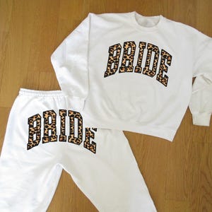 May include: White sweatshirt and sweatpants set. The word "BRIDE" is printed in a leopard print design across the chest of the sweatshirt and on the left leg of the sweatpants. The set is laid out on a wooden surface.