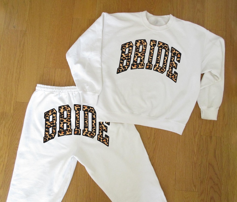 May include: White sweatshirt and sweatpants set. The word "BRIDE" is printed in a leopard print design across the chest of the sweatshirt and on the left leg of the sweatpants. The set is laid out on a wooden surface.