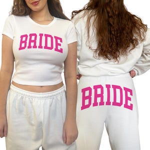 May include: White t-shirt and sweatpants set with the word "BRIDE" in pink block letters. The t-shirt is a cropped style, and the sweatpants have a drawstring waist. The set is designed for a bridal theme.