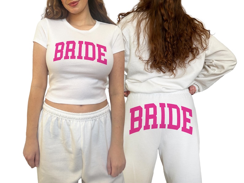 May include: White t-shirt and sweatpants set with the word "BRIDE" in pink block letters. The t-shirt is a cropped style, and the sweatpants have a drawstring waist. The set is designed for a bridal theme.