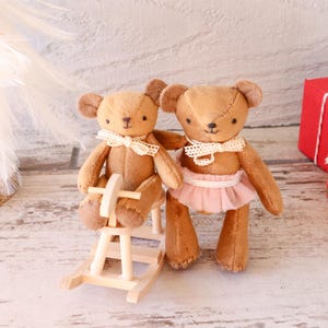 Puede incluir: Dos osos de peluche marrones con cuellos de encaje color crema. Uno está sentado en un pequeño balancín de madera, el otro lleva un tutú rosa. Los osos están hechos a mano, ideales para una habitación infantil.
