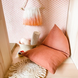 May include: A miniature dollhouse interior with a peach and white ombre wall hanging, a woven basket with rolled fabric, a peach pillow, and a woven jute placemat. The walls are decorated with a pink honeycomb pattern.