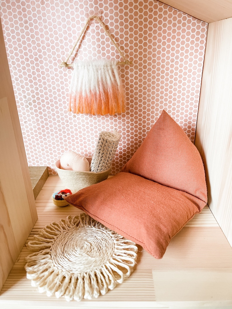 May include: A miniature dollhouse interior with a peach and white ombre wall hanging, a woven basket with rolled fabric, a peach pillow, and a woven jute placemat. The walls are decorated with a pink honeycomb pattern.