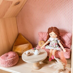 May include: A dollhouse interior with a doll seated on a pink cushion. The doll has red hair and wears a floral dress. A small wooden table holds a teapot and treats. A pink fuzzy ottoman and yellow beanbag are also present.