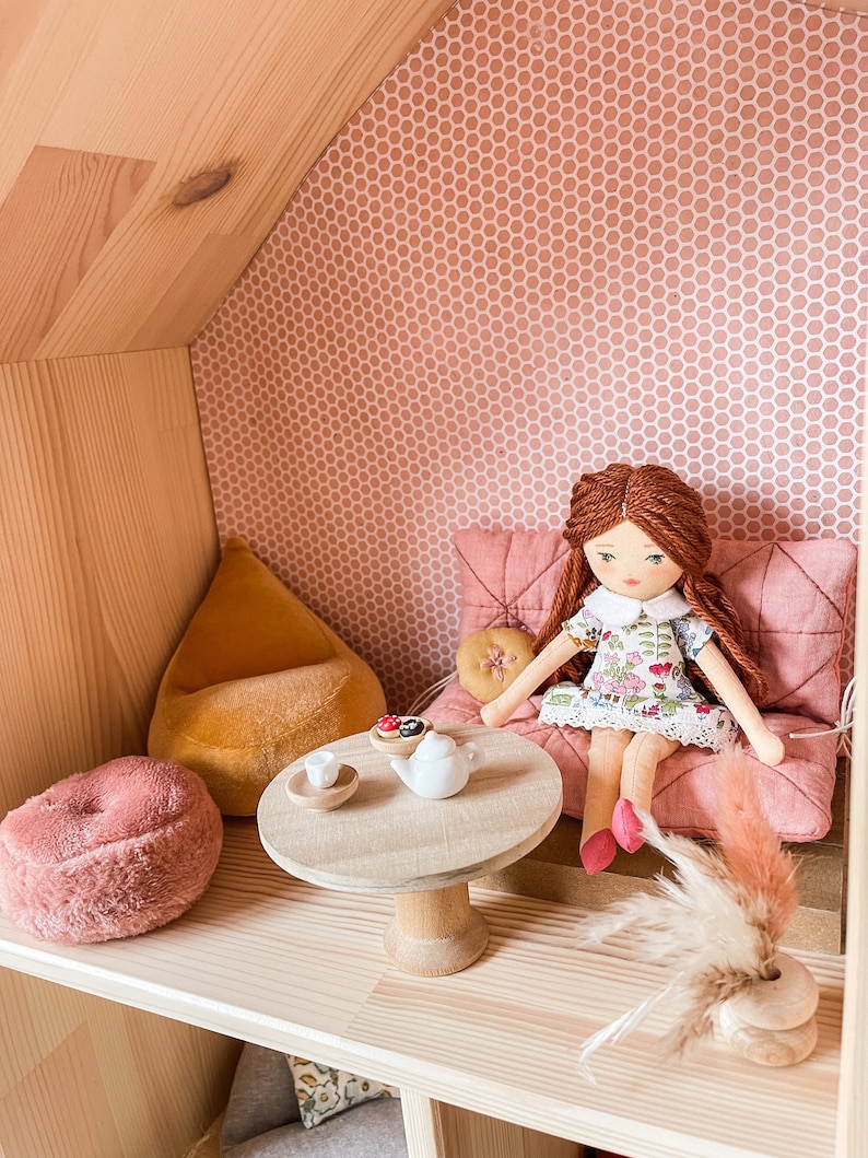 May include: A dollhouse interior with a doll seated on a pink cushion. The doll has red hair and wears a floral dress. A small wooden table holds a teapot and treats. A pink fuzzy ottoman and yellow beanbag are also present.