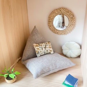 May include: A miniature dollhouse interior with a light gray pillow, a small floral pillow, and a white, round pillow. A small mirror with a woven frame hangs on the wall. A miniature plant and a stack of books complete the scene.
