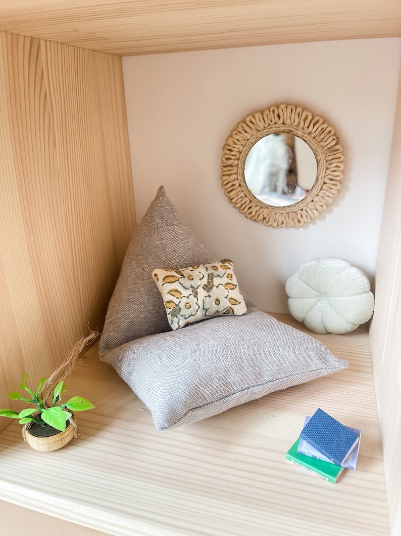 May include: A miniature dollhouse interior with a light gray pillow, a small floral pillow, and a white, round pillow. A small mirror with a woven frame hangs on the wall. A miniature plant and a stack of books complete the scene.
