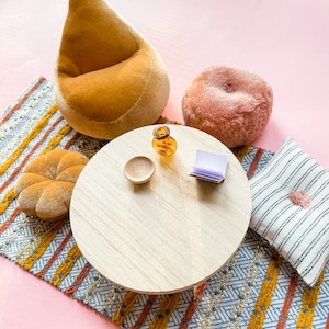 May include: A miniature living room scene with a round wooden table, a small orange glass bottle, and a small wooden bowl. A mustard-colored beanbag chair, a pink fluffy cushion, a patterned rug, and a striped pillow with a pink tassel complete the scene.