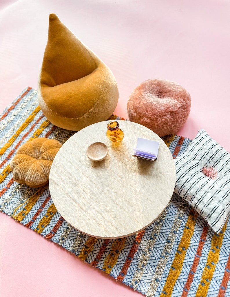 May include: A miniature living room scene with a round wooden table, a small orange glass bottle, and a small wooden bowl. A mustard-colored beanbag chair, a pink fluffy cushion, a patterned rug, and a striped pillow with a pink tassel complete the scene.
