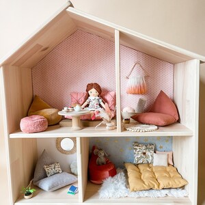 May include: A wooden dollhouse with multiple rooms, furnished with miniature pillows, beanbag chairs, and a doll. The dollhouse has a light wood finish and is decorated with patterned wallpaper and small accessories.