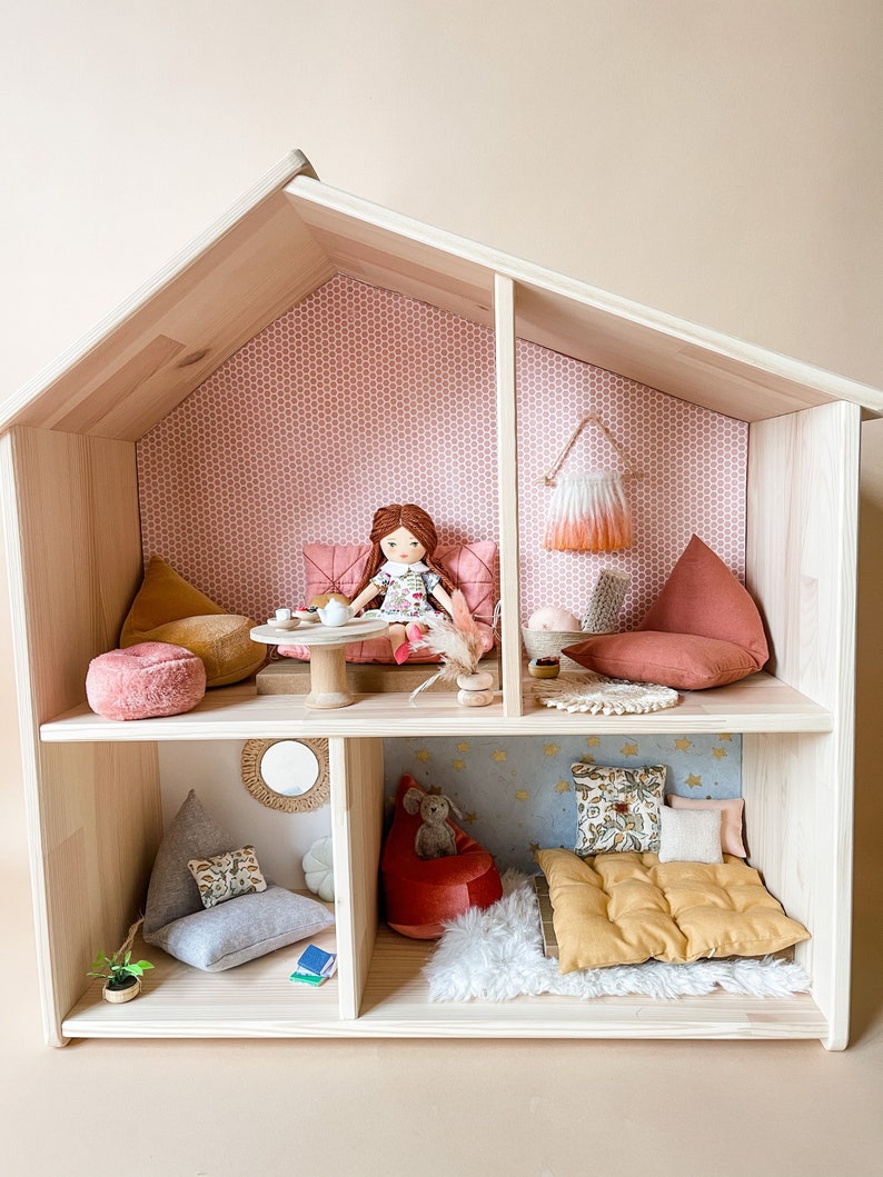 May include: A wooden dollhouse with multiple rooms, furnished with miniature pillows, beanbag chairs, and a doll. The dollhouse has a light wood finish and is decorated with patterned wallpaper and small accessories.