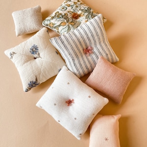 May include: An assortment of small, decorative pillows in various patterns and colors. The pillows include floral, striped, and solid designs, with colors like cream, pink, and green. Some pillows have decorative accents.