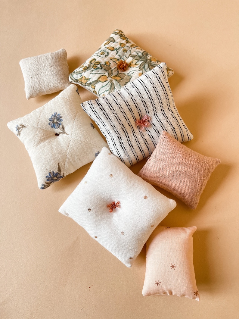 May include: An assortment of small, decorative pillows in various patterns and colors. The pillows include floral, striped, and solid designs, with colors like cream, pink, and green. Some pillows have decorative accents.