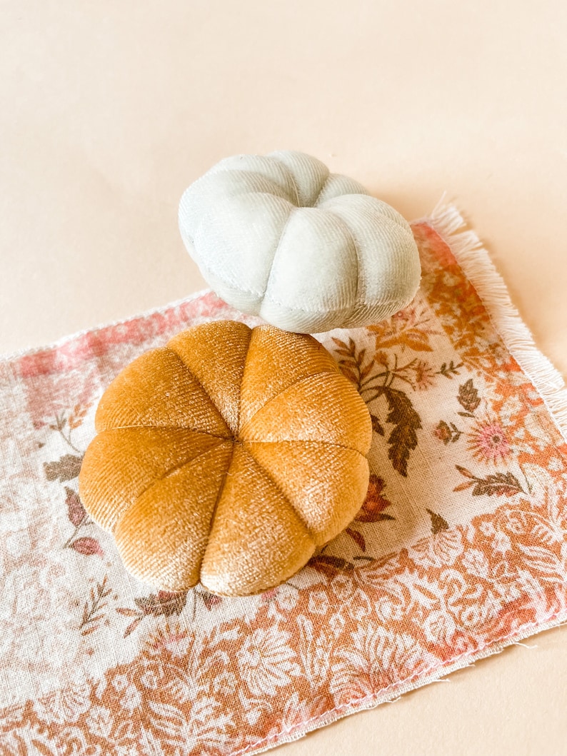 May include: Two plush, pumpkin-shaped decorations. One is a light sage green, the other is a warm golden yellow. They rest on a floral patterned fabric with a fringed edge, set against a neutral background. Decorative fall accents.