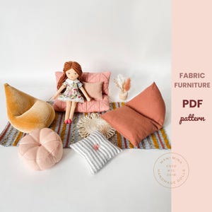 May include: A collection of miniature fabric furniture, including a velvet beanbag chair, a quilted pink cushion, and a striped pillow. A doll with red hair sits on a pink cushion. The image also includes a small rug and a decorative sun-shaped item.
