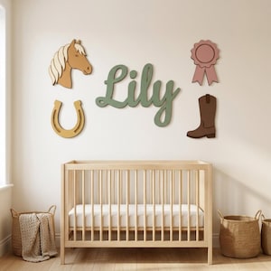 May include: A wooden cot sits below wall-mounted decor featuring the name Lily in green script, a tan horse head, a yellow horseshoe, a pink award ribbon, and a brown cowboy boot. The items are arranged on a plain wall above the light wood cot.