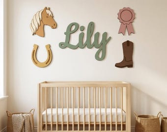 Puede incluir: Una cuna de madera se sitúa bajo decoraciones de pared que incluyen el nombre Lily en letra cursiva verde, una cabeza de caballo beige, una herradura amarilla, una escarapela rosa y una bota vaquera marrón sobre la pared.