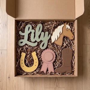 May include: A cardboard box filled with brown paper filler containing five wooden cutouts: a sage green script name sign reading Lily, a tan horse head, a yellow horseshoe, a dusty pink award ribbon, and a brown cowboy boot.