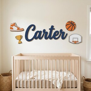 May include: A wall-mounted wooden cutout of the name Carter in dark blue script, accompanied by four separate wooden wall decorations: an orange and white high-top sneaker, a gold trophy, an orange basketball, and a white basketball hoop.