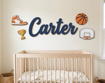 Puede incluir: Un recorte de madera montado en la pared con el nombre Carter en letra cursiva azul oscuro, acompañado de cuatro decoraciones de pared de madera separadas: una zapatilla alta naranja y blanca, un trofeo dorado, un balón de baloncesto naranja y una canasta blanca.
