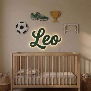 May include: A light-up wall sign displaying the name Leo in dark green cursive script, surrounded by wall-mounted cutouts of a soccer ball, a green soccer cleat, a gold trophy, and a white soccer goal frame above a wooden crib.