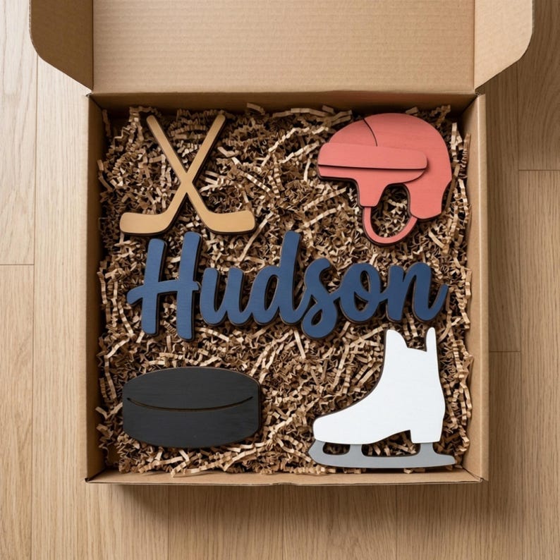 May include: A cardboard gift box filled with brown paper crinkle filler containing wooden cutouts: a tan hockey stick pair, a red hockey helmet, a dark blue script name cutout reading Hudson, a black hockey puck, and a white ice skate.