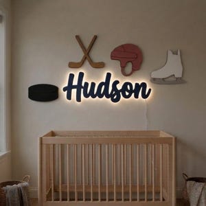 May include: A wall-mounted LED sign displaying the name Hudson in dark blue script, illuminated from behind. Surrounding the name are wooden cutouts of two crossed hockey sticks, a red hockey helmet, a white ice skate, and a black puck.