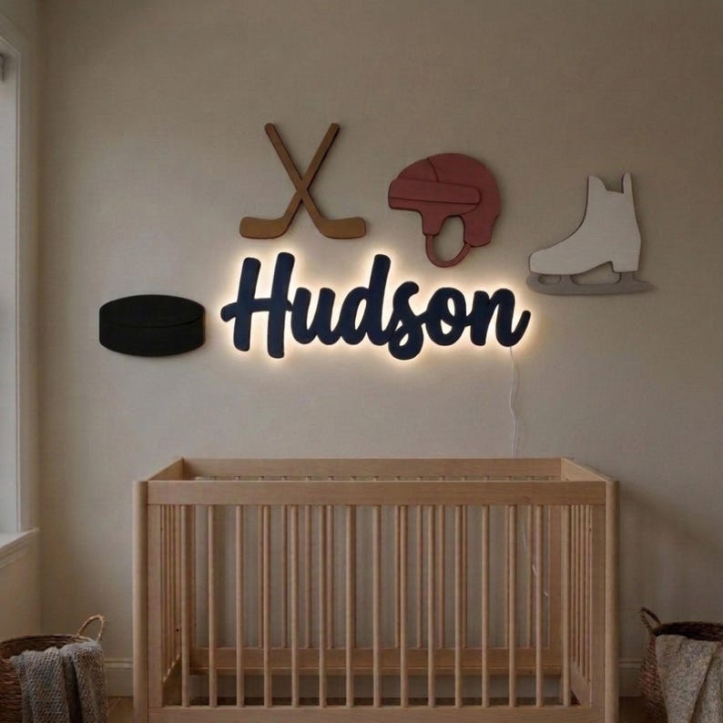 May include: A wall-mounted LED sign displaying the name Hudson in dark blue script, illuminated from behind. Surrounding the name are wooden cutouts of two crossed hockey sticks, a red hockey helmet, a white ice skate, and a black puck.