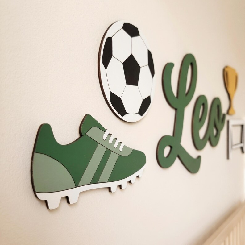 May include: A wall-mounted set featuring a green soccer cleat, a black and white soccer ball, the name Leo in green cursive script, and a gold trophy icon. These decorative cutouts are arranged to create a sports-themed display.
