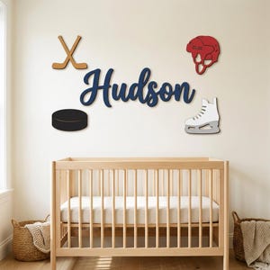 May include: A wall decor set featuring the name Hudson in dark blue script, accompanied by a tan wooden hockey stick cross, a red hockey helmet, a black hockey puck, and a white ice skate, all arranged above a light wood cot.