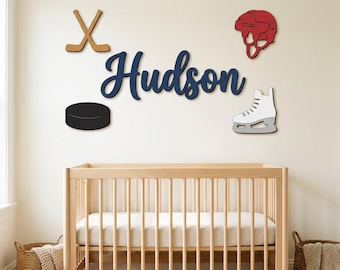Puede incluir: Un conjunto de decoración de pared con el nombre Hudson en letra cursiva azul oscuro, acompañado de palos de hockey de madera beige cruzados, un casco rojo, un disco negro y un patín de hielo blanco sobre una cuna de madera clara.