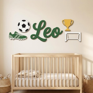 May include: A wall decor set featuring a black and white football, a green football boot, the name Leo in dark green cursive script, a gold trophy, and a white football goal outline, all arranged horizontally above a wooden cot.