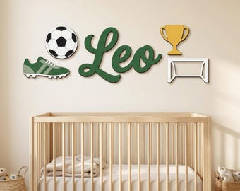 Puede incluir: Un conjunto de decoración de pared que incluye un balón de fútbol blanco y negro, una bota de fútbol verde, el nombre Leo en letra cursiva verde oscuro, un trofeo dorado y el contorno de una portería de fútbol blanca.