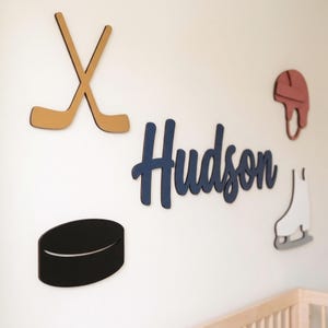 May include: A set of wall-mounted wooden cutouts featuring the name Hudson in dark blue script, two crossed tan hockey sticks, a black hockey puck, a red hockey helmet, and a white ice skate with a grey blade, all arranged on a plain wall.