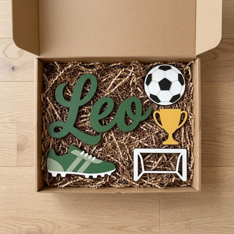 May include: A cardboard gift box filled with brown paper filler containing a dark green wooden cutout of the name Leo, a black and white soccer ball, a yellow trophy, a green soccer cleat, and a white soccer goal frame cutout.