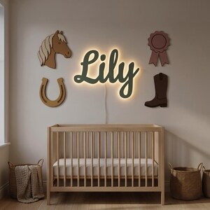 May include: A wall-mounted LED sign displaying the name Lily in dark green script with a warm backlight. Surrounding the sign are four wooden wall decorations: a brown horse head, a tan horseshoe, a dark brown cowboy boot, and a mauve ribbon.