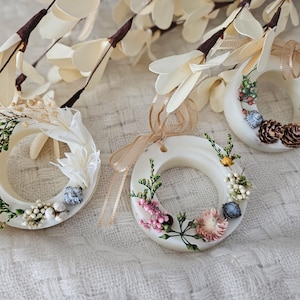 May include: Three white wax candle rings decorated with dried flowers and greenery. The rings are tied with brown ribbon and have a rustic, natural aesthetic.
