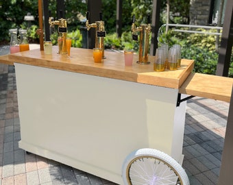 Può includere: Un carrello bar mobile con base color crema e piano d'appoggio in legno chiaro. Presenta tre rubinetti per la birra color oro, bicchieri e una ruota di bicicletta bianca. Il carrello è progettato per eventi e riunioni all'aperto.