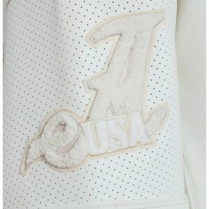 May include: A white perforated jacket sleeve featuring a large, textured, cream-colored applique patch. The patch displays a stylized letter A with the letters USA integrated into the design, rendered in a fuzzy, plush material with stitched edges.