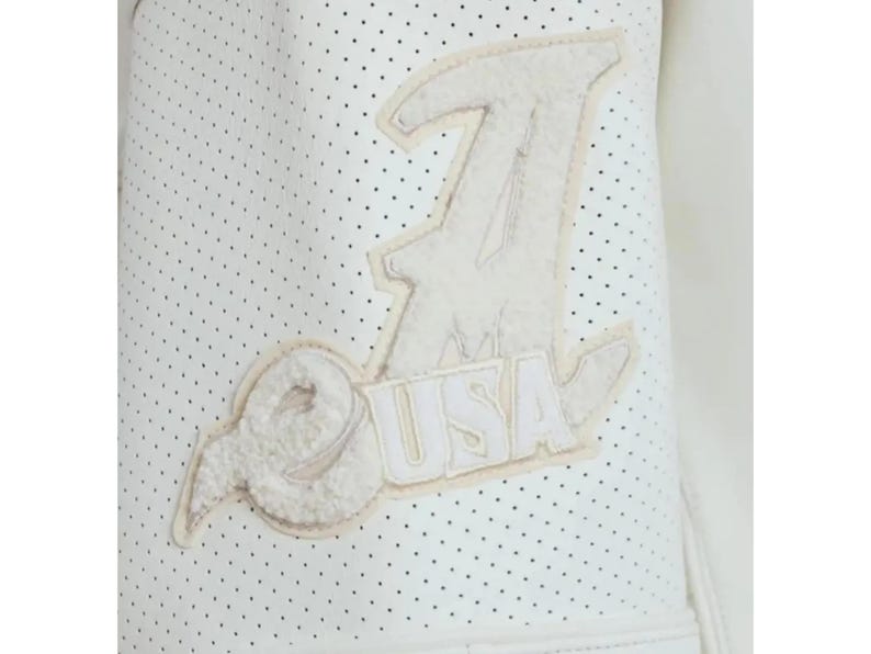 May include: A white perforated jacket sleeve featuring a large, textured, cream-colored applique patch. The patch displays a stylized letter A with the letters USA integrated into the design, rendered in a fuzzy, plush material with stitched edges.