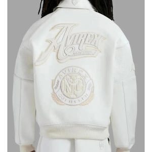 May include: A white varsity-style jacket featuring a large embroidered Avirex New York logo across the upper back and a circular Avirex Gold Club NYC emblem below. The jacket has a collared neckline, ribbed hem, and textured sleeve panels.