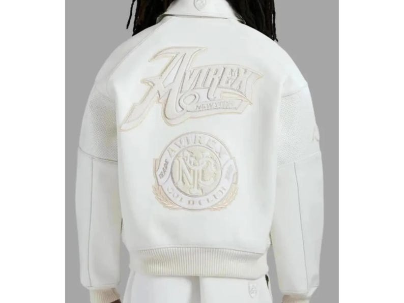 May include: A white varsity-style jacket featuring a large embroidered Avirex New York logo across the upper back and a circular Avirex Gold Club NYC emblem below. The jacket has a collared neckline, ribbed hem, and textured sleeve panels.