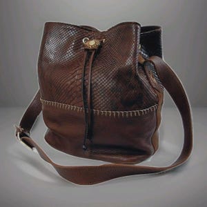 May include: Brown leather bucket bag with a snakeskin texture on the upper portion and a smooth leather base. The bag features a drawstring closure, a long adjustable strap, and a decorative clasp.
