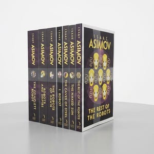 Può includere: Un cofanetto di libri di Isaac Asimov, con titoli come "The Complete Robot" e "The Rest of the Robots". I dorsi sono scuri con scritte dorate, e la scatola presenta un design viola e oro con illustrazioni di robot. Una collezione di fantascienza.