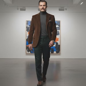 以下が含まれることがあります： 茶色のコーデュロイブレザー、グレーのタートルネック、濃い緑色のパンツを着た男性。 ブレザーにはポケットチーフが付いています。 男性は時計と茶色の革靴を履いています。 背景には抽象的な絵画があります。