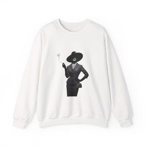 May include: White sweatshirt featuring a black and white graphic of a woman in a suit and wide-brimmed hat, smoking a cigarette. The image is a stylized illustration, with the woman's face partially obscured. The sweatshirt has a crew neck and long sleeves.