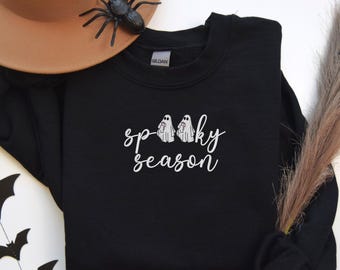 May include: Black sweatshirt with the words "spooky season" in white script, along with two small ghost illustrations. A brown hat and a plastic spider are in the background, suggesting a Halloween theme.