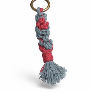 Könnte beinhalten: Ein handgefertigter Makramee-Schlüsselanhänger mit einem Messingring. Der Schlüsselanhänger hat ein gewebtes Design in Grau und Rot mit einer grauen Quaste am unteren Ende. Dieses Accessoire ist perfekt, um Ihren Schlüsseln oder Ihrer Tasche einen Hauch von Bohème-Stil zu verleihen.