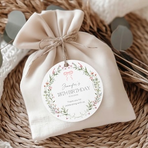 May include: A cream-coloured fabric gift bag tied with string, with a circular tag. The tag has a floral wreath, a pink bow, and the text "Jennifer's 18th Birthday 10.22.2025". The tag also includes the text "Thank you for celebrating with us."